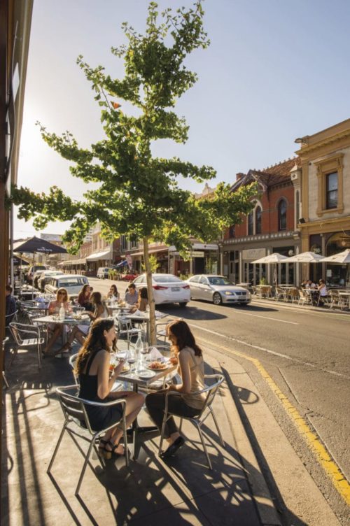 2 women drinking and eating on a terrace in a busy street with a blue sky in Adelaide.